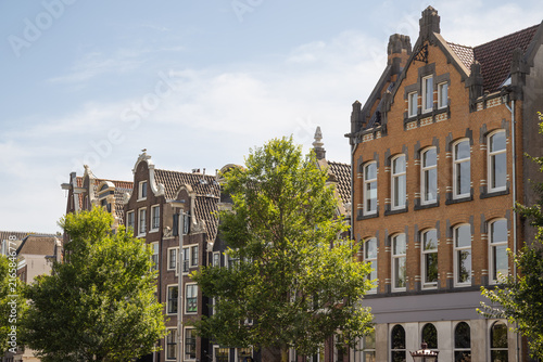 Classic Dutch architecture featuring historical brick house facades with ornate roof gables next to lush green trees against a partly cloudy summer sky in Amsterdam