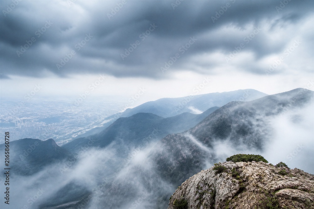 Obraz premium Stormy clouds loom over mist-shrouded mountains. Rocky peak juts from fog, crowned with sparse green life. Distant city sprawls beneath rolling hills. Mist curls between peaks, soft and silent