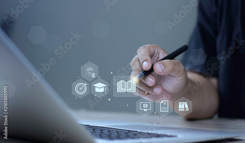 Konstfotografi Online course training, digital knowledge database, and skill development concept with hand using stylus pen pointing at glowing book and e-learning HUD icons on laptop screen