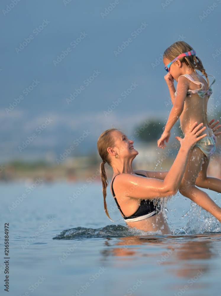 Fototapeta premium Woman in a black swimsuit lifts her small daughter high above the calm sea, the child laughing with her arms out against the sky