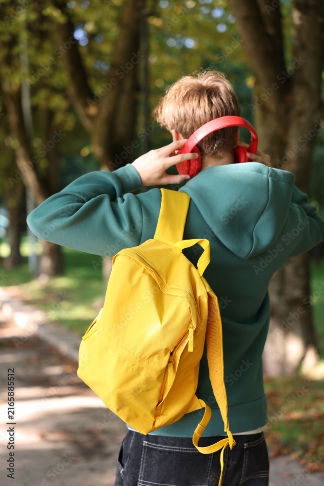 Fototapeta premium Student with yellow backpack putting on headphones in park on sunny day, back view
