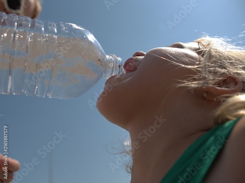 Obraz na plátně Blonde girl in a green top drinks water from a plastic bottle held above her hea