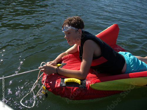 Картината върху платно Teenage boy in a black vest lies on a red inflatable and coils the tow rope in h