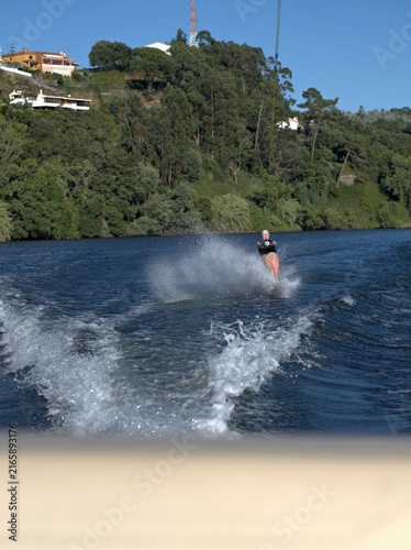 Bild auf Leinwand Water skier throws up a curtain of spray on a calm river, hillside houses and de