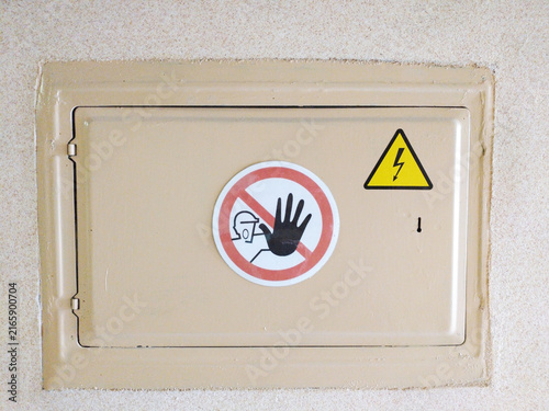 An electrical distribution box on a light-colored wall. Warning signs are displayed on the door, indicating the danger of electric voltage and the need to observe safety rules.