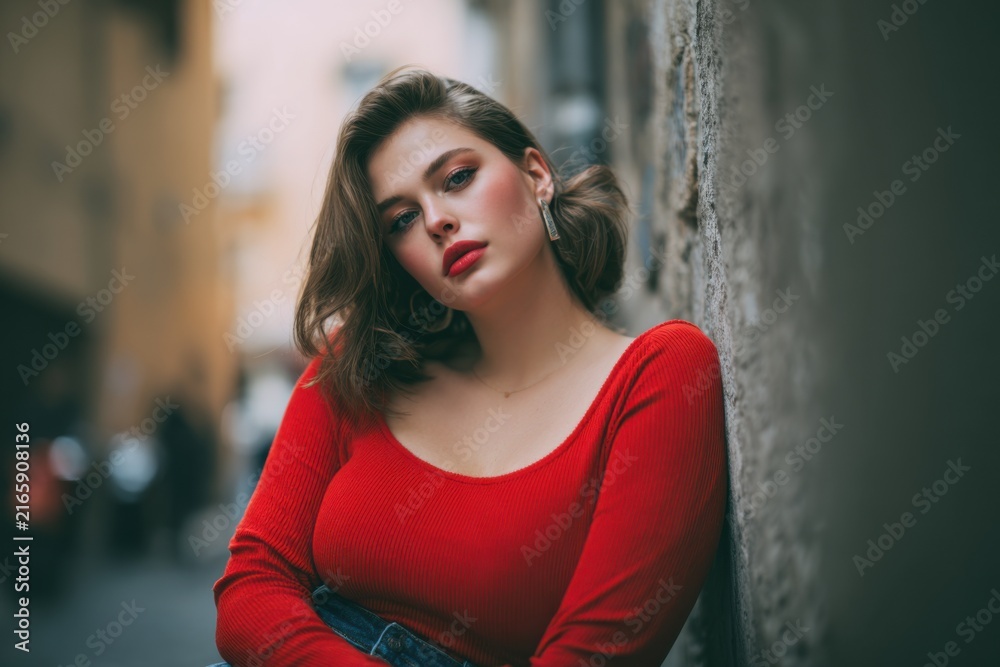Fototapeta premium Curvy model in red top leaning against alley wall, confident street portrait with soft bokeh background, bold makeup, elegant pose and fashionable outfit conveying attitude and evening glamour