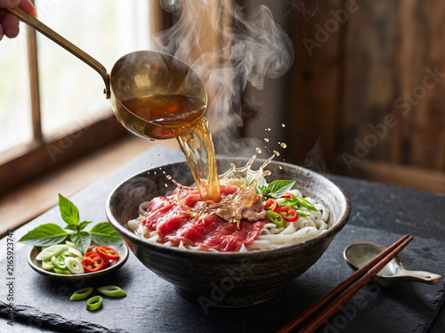 Dynamic action shot of pouring hot steaming broth over raw beef pho noodles with splash effect