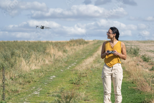 Woman holding a remote controller and piloting a flying drone along a countryside path surrounded by tall grass under a cloudy sky.