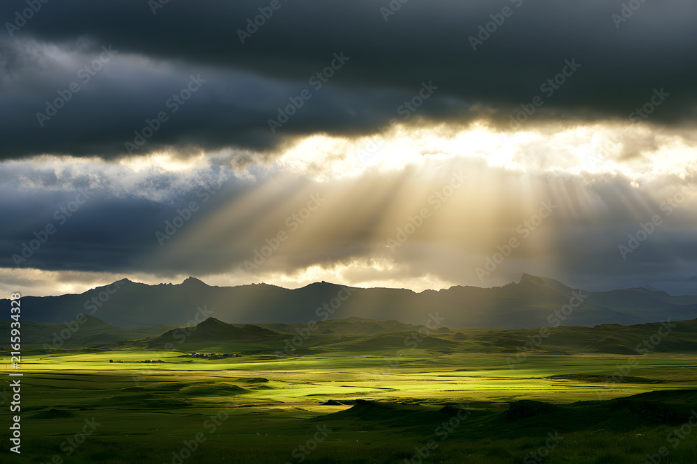 Fototapeta premium Sunlight penetrates through gray clouds above green Icelandic pastures, mountains. Golden rays illuminate grassy fields beneath a cloudy sky at sunrise. A picturesque rural scene displays striking wea