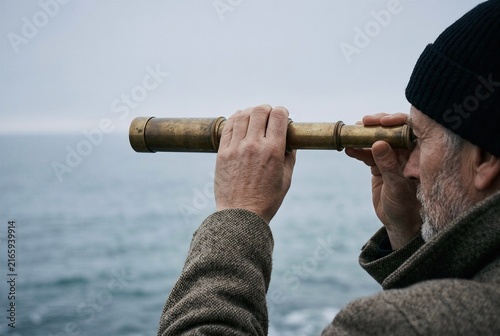 Faceless over the shoulder view of a hand holding a blank brass spyglass toward the sea. Cinematic haze for maritime exploration concepts.