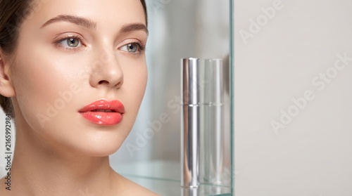 Young woman with glowing skin and glossy lips posing with cosmetic product bottle.