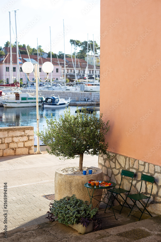 Fototapeta premium Outdoor mediterranean breakfast setup with olive tree in pot, coffee, croissants, and orange juice on a bistro table overlooking Sali marina in Croatia.