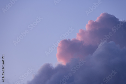 Fluffy cumulus cloud formation in a clear sky at sunset. The cloud has soft pink and purple highlights from the sun, contrasting with deep grey-blue shadow. Perfect for background design.