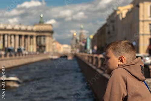 Quadro su tela A cute boy in a warm brown sweatshirt standing on a bridge and thoughtfully looking at the river