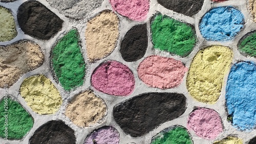 Close-up of a decorative stone wall surface featuring colorful painted rocks in various vibrant hues. Concept of urban landscaping, creative architecture, and abstract texture pattern background.