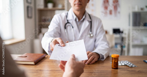 Doctor handing prescription form to patient during medical office consultation with medicine bottle and pills on wooden desk in bright clinic setting professional health care