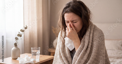 Sick young woman suffering from seasonal flu sitting in bedroom wrapped in a warm knitted beige blanket holding tissue paper while feeling bad during illness