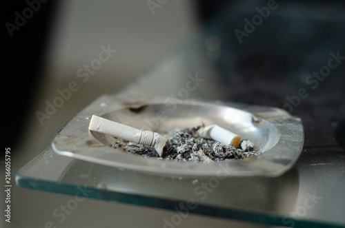 Fotografía Two Cigarette Butts and Ash in a Metal Ashtray on a Glass Surface