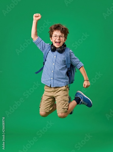 Happy energetic school boy jumping high with backpack and headphones on green background.