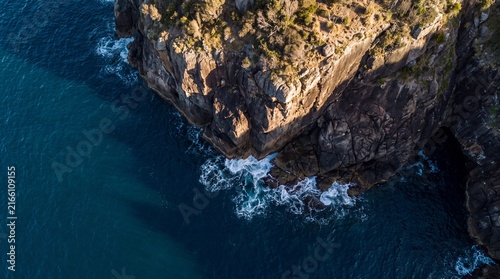 Dramatic coastal cliff aerial view, rugged ocean rock formation with crashing waves, steep seaside precipice at golden hour