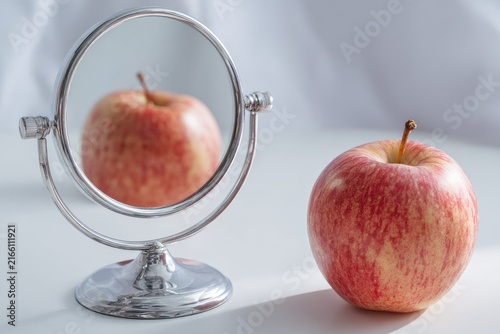 A small round silver mirror reflects a red apple, juxtaposed with an identical apple
