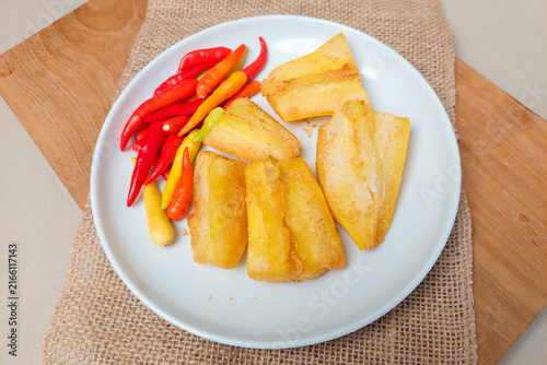 Obraz na plátně Fried cassava pieces served on a white plate with red chilies and a rustic-style natural garnish, set against a wooden background