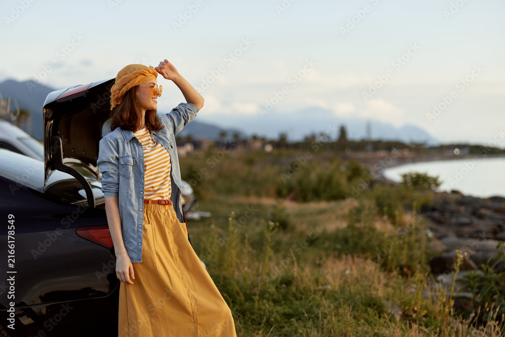 Naklejka premium Woman outdoors by a car on a coastal landscape, wearing a denim jacket and mustard dress, sunlit moment, joyful pose, gentle breeze, vibrant colors, relaxed lifestyle