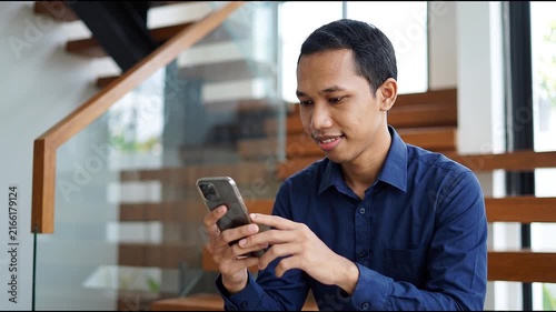 Smiling happy young Indonesian man holding cell mobile phone using smartphone sitting at home on couch, scrolling social media, checking financial apps, buying online, texting messages. 