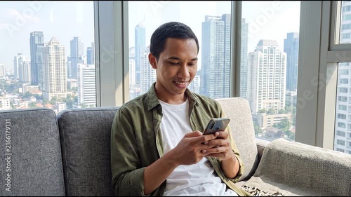 Smiling happy young Indonesian man holding cell mobile phone using smartphone sitting at home on couch, scrolling social media, checking financial apps, buying online, texting messages. 
