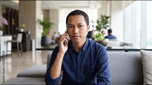 Smiling happy young Indonesian man holding cell mobile phone using smartphone sitting at home on couch, scrolling social media, checking financial apps, buying online, texting messages. 