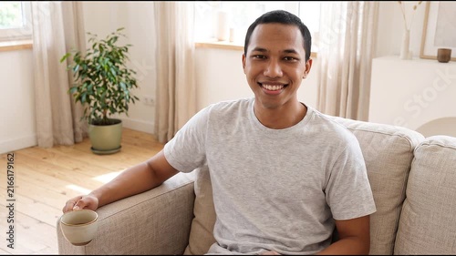 Smiling happy young Indonesian man holding cell mobile phone using smartphone sitting at home on couch, scrolling social media, checking financial apps, buying online, texting messages. 