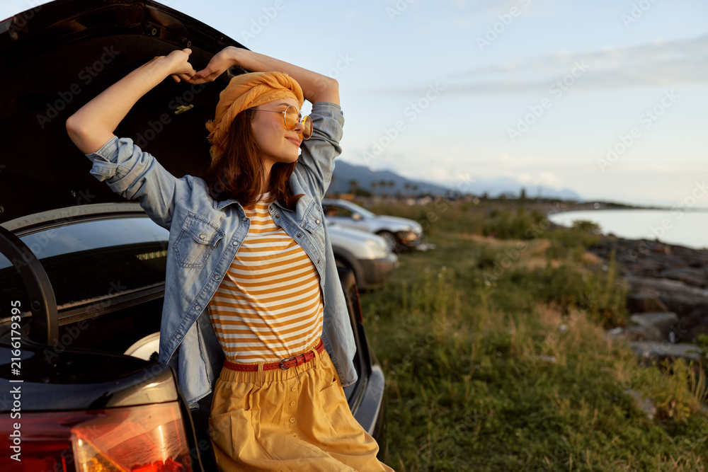 Fototapeta premium Cheerful woman outdoors, leaning against a car trunk, smiling in sunlight, striped top and denim shirt, relaxed lifestyle, gentle breeze, natural tones, coastal scenery
