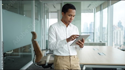 Busy young Indonesian business man executive standing in office using digital tablet. Young professional businessman corporate manager wearing suit and glasses holding tab working on finance project