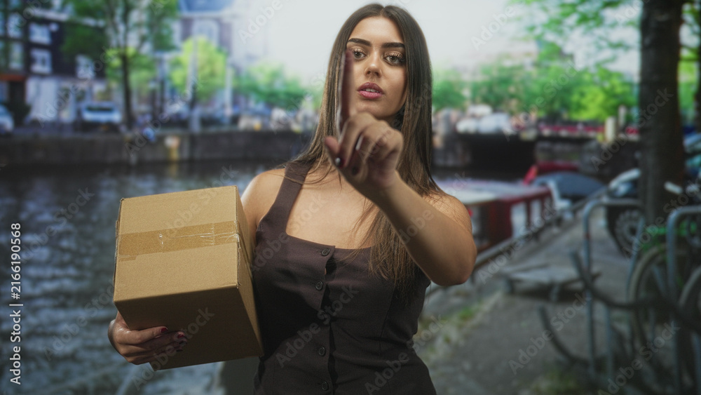 Fototapeta premium Woman holding cardboard box points index finger on street by a canal and bicycles; determination delivery.