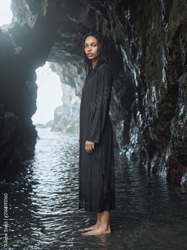 Woman in dark robe standing in sea cave, mystical light