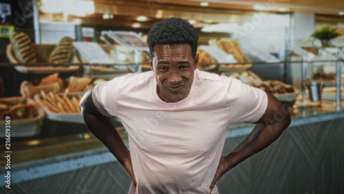 Bild auf Leinwand Man in pink tshirt clutching stomach with hands amid bakery pastries by bread display at building counter; pain nausea urgency