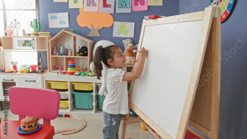 Foto Girl drawing on a whiteboard easel in a preschool classroom with toys and storage bins; concentration creativity learning