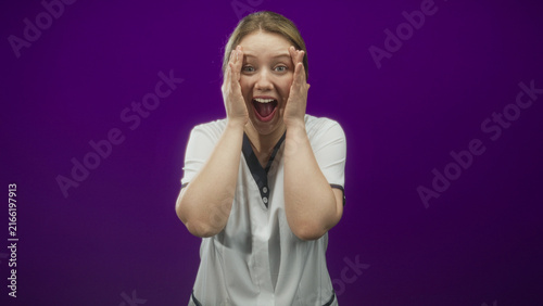 Quadro su tela Woman in white nurse uniform pressing hands to face with mouth open and wide eyes in studio purple backdrop; surprise celebration