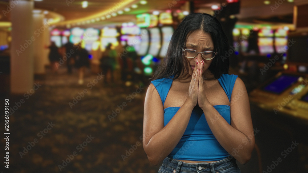 Fototapeta premium Woman with hands pressed together in prayer in a casino with slot machines and blurred patrons; desperation hope loss.