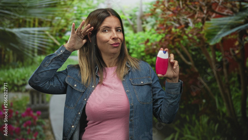 Quadro su tela Middle aged hispanic woman holding pink toothpaste bottle, hand to head for toothache in forest setting with garden path visible; annoyance hesitation