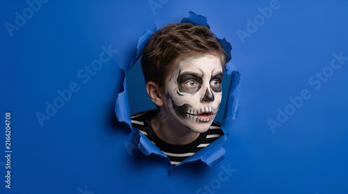 Boy with skull face paint peeking through torn blue paper. Halloween costume, spooky theme, creative concept.
