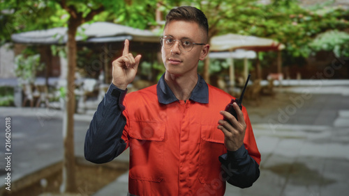 фотографія Young blond man in orange coverall holding a walkietalkie and points finger up on a street cafe terrace while wearing glasses and looking focused; alert readiness