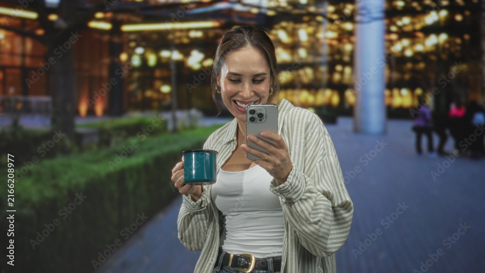 Fototapeta premium Woman holding smartphone and teal mug, smiling while reading a message on her phone outside illuminated building entrance; joy connection.