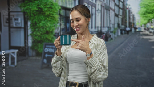 Принти на полотні Woman holds a teal mug and points finger on a street outside a cafe while smiling and gesturing to camera; cheerful confidence