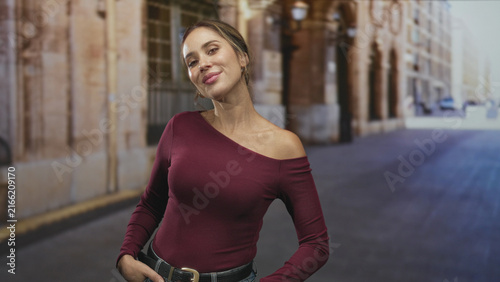 Quadro su tela Woman smiling with one shoulder exposed and hands on hips on a street with stone arches and pavement; confidence approachable