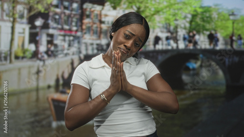 Fotografía Woman wearing white polo shirt with hands clasped in prayer and pleading facial expression on a street beside a canal bridge; pleading vulnerability