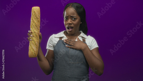 Konstfotografi Baker woman holding long baguette in one hand with hand on chest and mouth open in studio; surprise concern