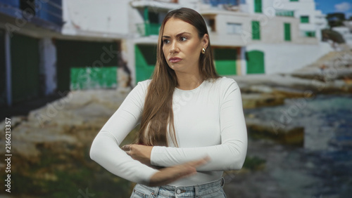 Photography Woman arms crossed and pursed lips standing before a white coastal building with green shutters and visible jeans waist; frustration doubt