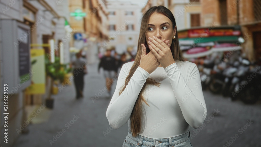 Fototapeta premium Woman covering mouth with hands and long hair on a crowded street with scooters and shops; surprise reaction.