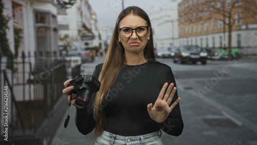 Obraz na płótnie Woman holding a dslr camera, palm raised in a stop gesture on a street with cars and buildings, wearing glasses and black top; disgust